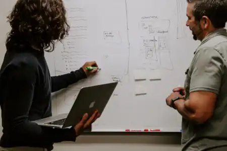 A woman draws on a whiteboard while a man in a polo shirt looks on