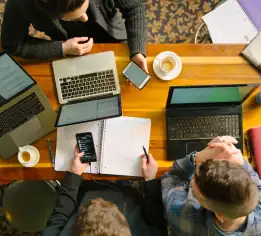 Several dudes gather around a table crowded with laptops and notebooks